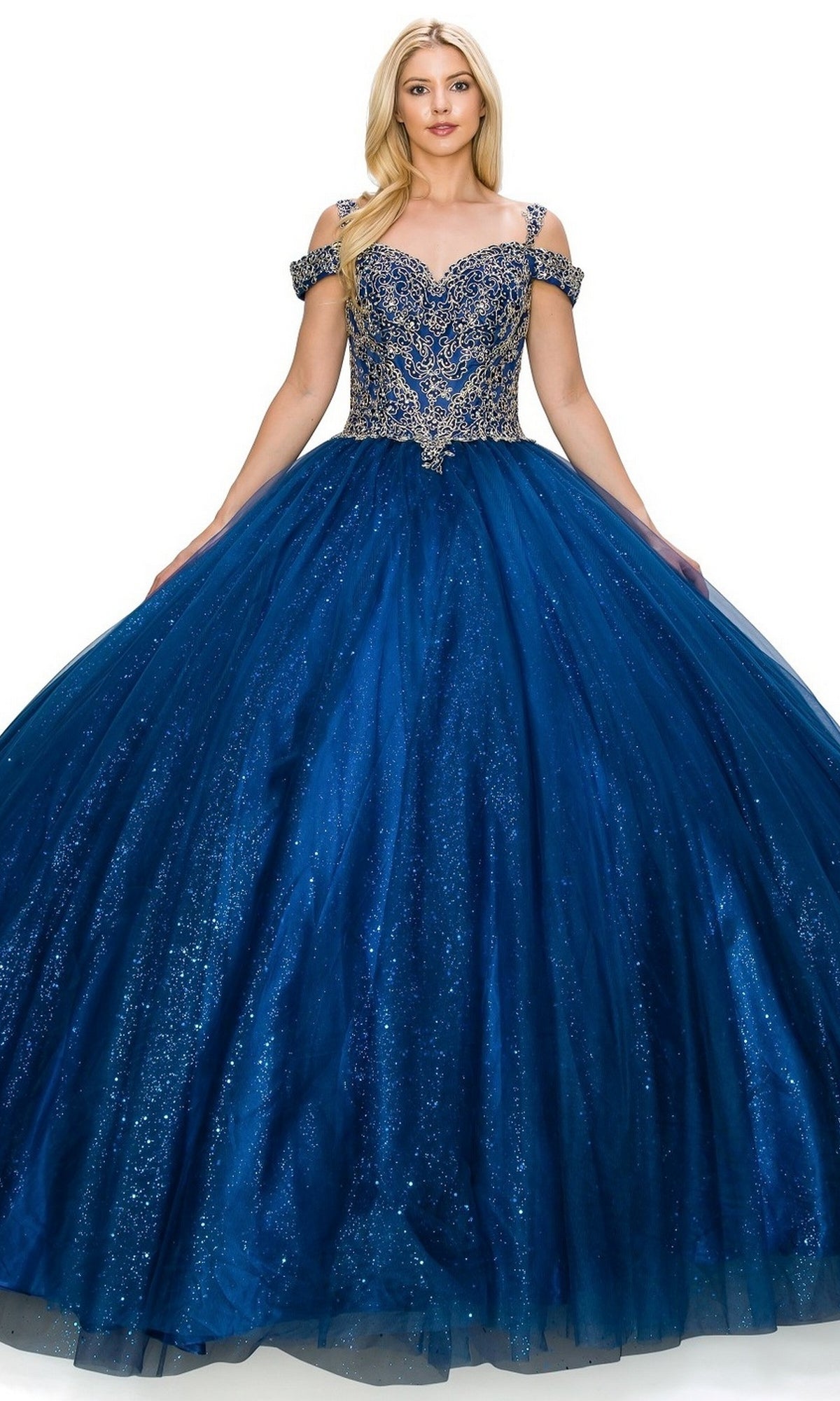 A woman stands against a white background wearing the Quinceanera Ball Gown 8028J, a sparkling royal blue off-the-shoulder dress featuring ornate silver embroidery on the bodice and straps.